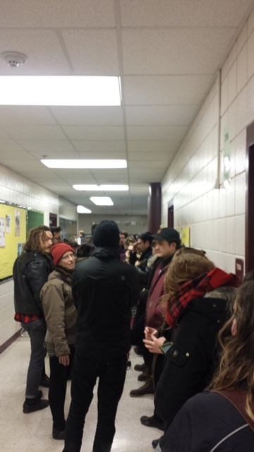 An industrial looking school hallway with fluorescent lighting. A group of people lean up against one wall and wait for the line to advance.