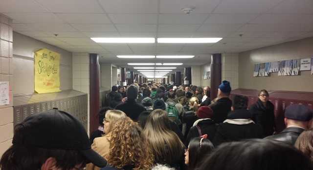 A large group of humanity swells from directly in front of the camera to all the way down to the end of a long school hallway.