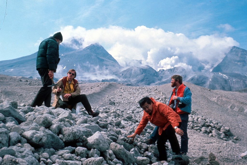 Image shows four geologists gathered on a jumbled pile of pumice. Two of them on the left are talking to each other. One on the right is watching those two, while the fourth man is leaning down to grab a bit of pumice. In the background, Mount St. Helens sits in clouds of steam.