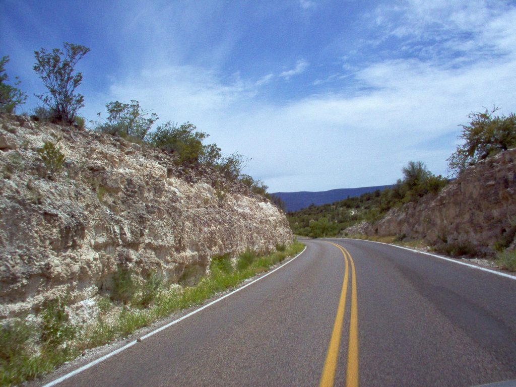 Image shows a two-lane road curving gently through low white limestone cliffs. The cliffs are covered in scrubby brush. There are some thin white clouds on the horizon and brilliant blue skies overhead.