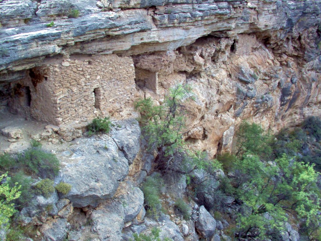 Image is a close view of a cliff. The top is layered limestone. There is a long cave where a softer layer eroded back: within that overhang are small buildings built of limestone blocks, with tiny openings for doors and windows. There is a large building at the center-left, with smaller buildings wedged in between blocks of solid stone beside it. The smaller rooms may be storerooms. Below the ruins, thick gray blocks of travertine form the cliff. There are small trees clinging to the cliff.
