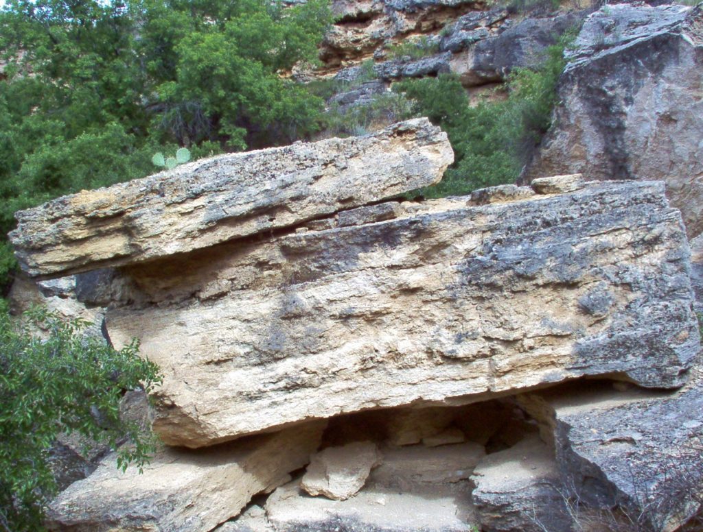 Image shows a stack of travertine blocks. They are a mottled gray and tan color, with the tan showing where the surface has been knocked off. The travertine is layered but massive. There is a thinner slice balanced on top of the bulky block, which in turn is resting on a jumble of smaller stones.