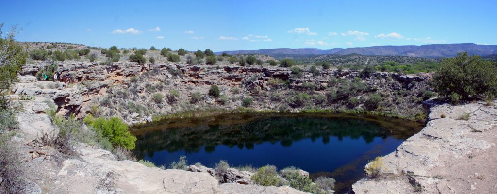 Image is a panorama overlooking a sinkhole. The water is reflecting the magnificent blue sky and the cliffs rimming it. The cliffs are grayish-white limestone, dotted with brushy trees, and filled with tiny caves. A range of mountains is visible in the distance.