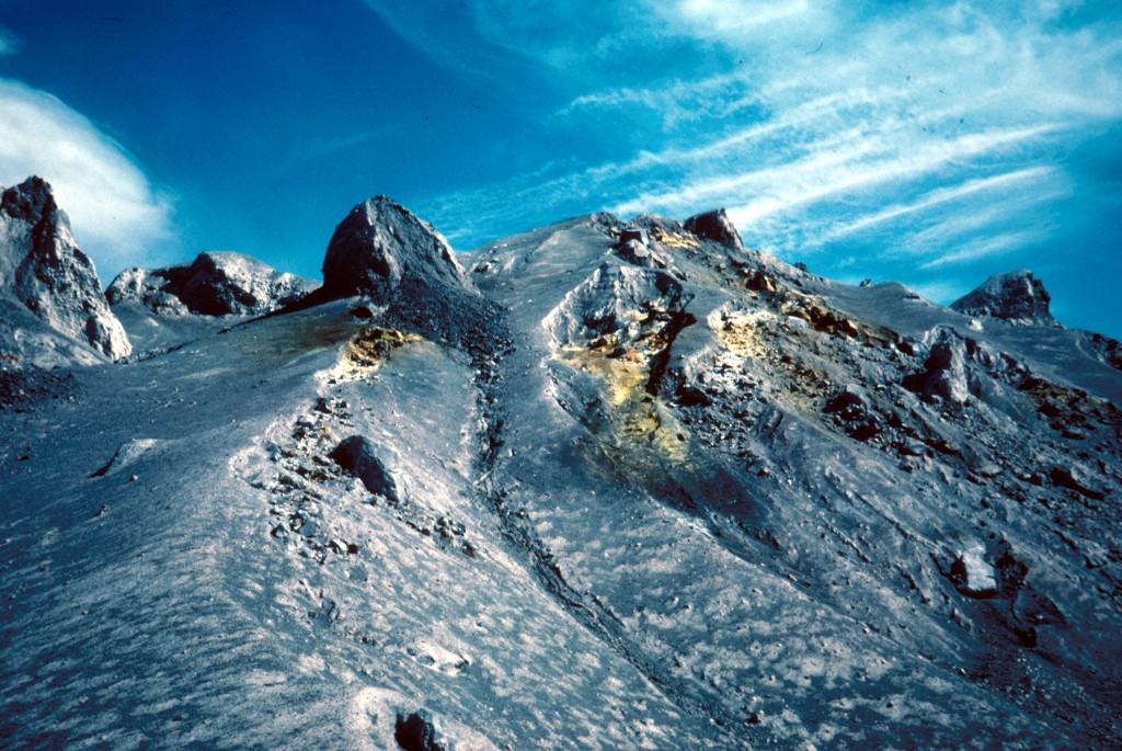 Image is taken from the base of a steep slope of mottled gray and light gray volcanic deposits, rising to a crest with boulders sticking out. Erosion has begun to cut shallow ravines int he slopes. Near the top, there are patches of harvest yellow where fumaroles have deposited sulfur: the color really pops against all that gray. The sky is deep blue with a few wispy white clouds.