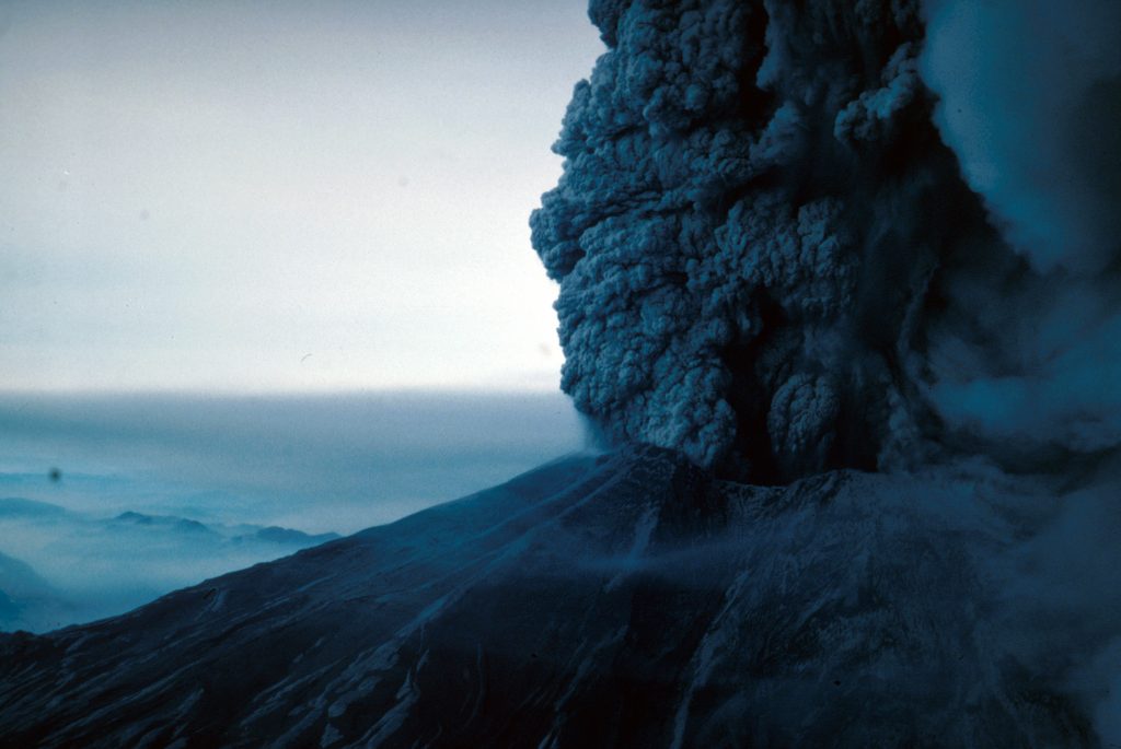 Image shows the upper part of Mount St. Helens's truncated summit, with an enormous ash cloud roaring from it. Image is in color: the cone and cloud are dark blue-gray. The valley in the distance is covered in pale gray ash.