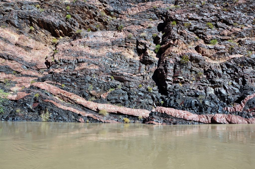 Image shows the Vishnu Schist, a black rock with veins of pink streaked through it. The Colorado River is visible at bottom - here, it is a muddy gray-green, with just a faint reflection of the rock wall above it.