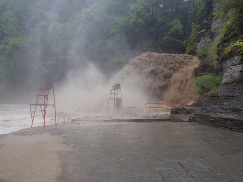 Image shows a sediment-laden torrent of water plunging over a cliff and roaring through a lake. Mist from the churning water is engulfing some metal lifeguard chairs. Muddy water has risen and is starting to cover the paved walkway that's normally high and dry.