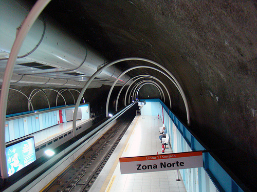 Image shows a Metro platform, looking down from near the ceiling. The ceiling is an arch of gray-brown rock. There are rails and ventilation equipment toward the left. On the right is a sign saying Zona Norte, and a platform with people sitting on benches against a wall. Everything is brightly lit.