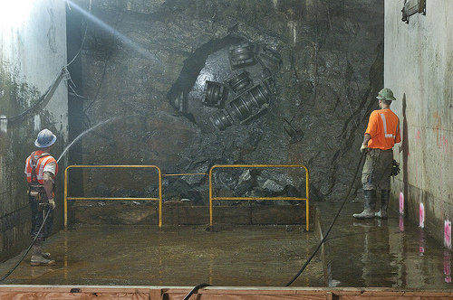 Image shows a tunnel boring machine cutting through a rock wall. There's a hole in the middle of the brownish-gray rock where the machine is chewing through. Portions of the machine are visible: it's metal darker than the rock, with several rotating bits. There are men in orange safety vests standing to either side, watching the machine work.