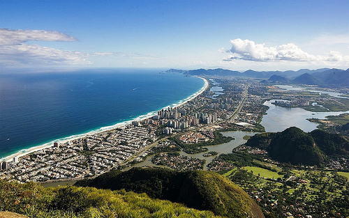 Image shows a long, curving spit of land between the deep blue waters of a bay and the shallow, brownish waters of a lagoon. Mountains are visible in the foreground and background. The flat land is filled with buildings.