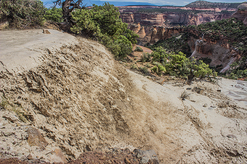 Image shows a torrent of muddy water plunging over a hillside. In the background, the image overlooks red rock canyons, pinon and juniper forests, and several cataracts unleashed by the rains.