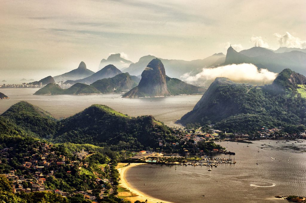 Image shows the iconic mountains and beaches of Rio. Sugarloaf and Corcovado are in the center right. There are some low clouds skimming the peaks, and a pinkish haze in the air, as if it's sunrise or sunset. The beaches glow a golden-white.