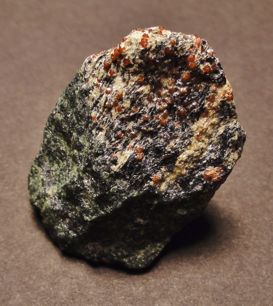 Image shows a small, oblong piece of rock standing on a black surface. The rock has glossy green minerals plastered to its left side. It shades up into dark navy blue streaked with golden colors. The top is studded with tiny red garnets.