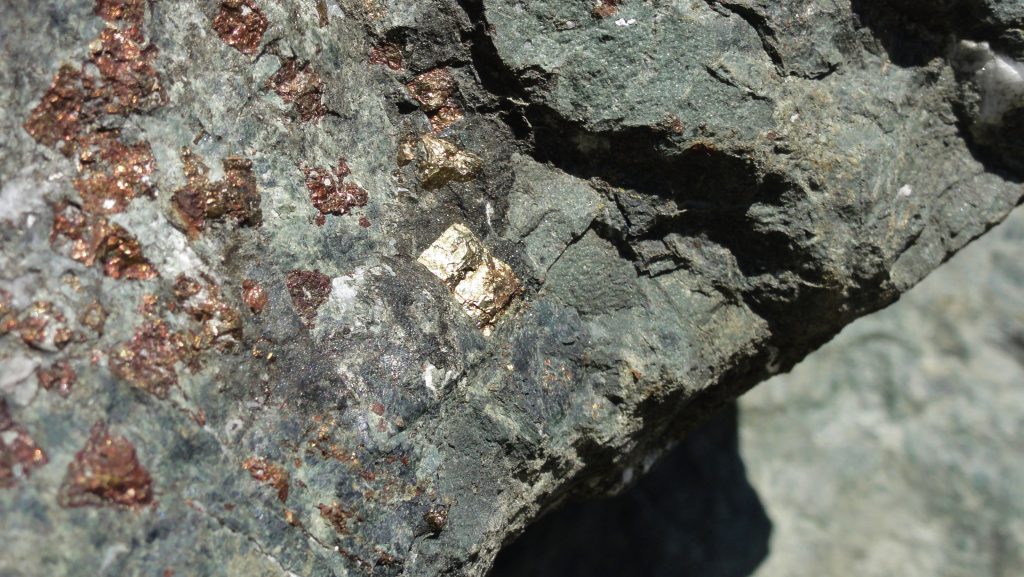 Image shows a slab of massive blueschist. Most of the minerals are too small to see with the naked eye in this one, giving it an overall greenish-blue appearance. There is a large square pyrite in it, and flecks of pyrite throughout it.