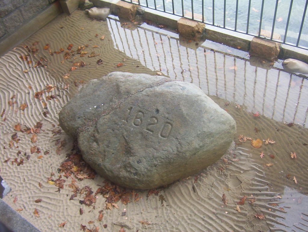 Image shows the piece of Plymouth Rock kept at Plymouth, Mass. It's a small, nondescript gray granitic boulder with the number 1620 carved into it. A seam runs through one side, filled with a coarse material. Patterns are raked into the sand around it. The grating that keeps people from taking more bits of it can be seen in the background.