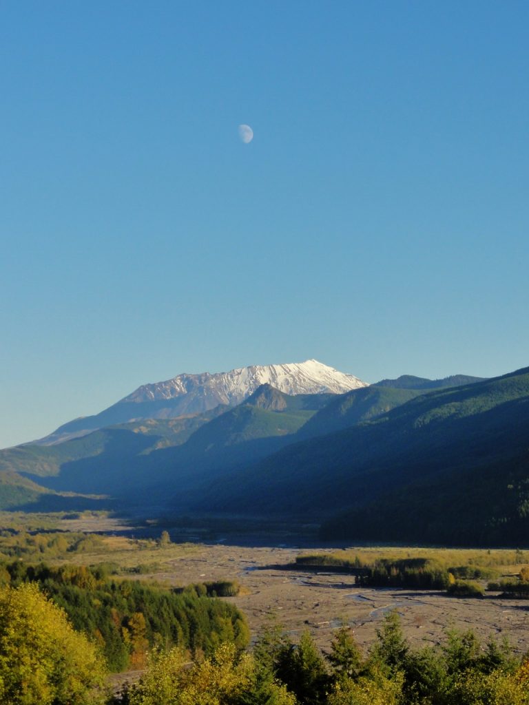 Image shows Mount St. Helens, snow-capped, rising over the Toutle River Valley near sunset. The moon is nearly full in the blue sky above the volcano.