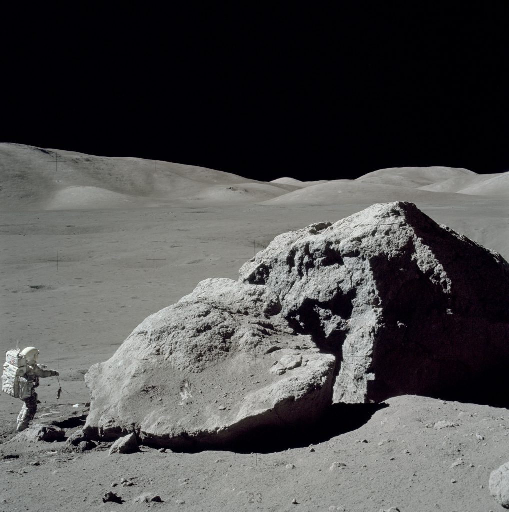 Image shows a man in a space suit standing in front of a huge, broken rock that's roughly the size of a house. They're on something of a plain, with low rolling hills standing pale gray against the black sky beyond.