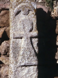 Photo of a symbol carved in relief on a small stone pillar. A rough skirted stick figure under a crescent and circle.