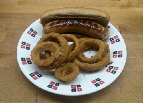 Photo of brat in wheat bun with onion rings on the same wooden counter.