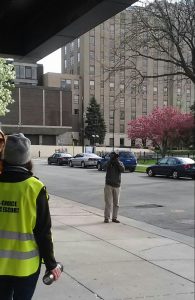 Photo of man standing on the sidewalk with a DSLR camera and longer lens, with camera pointed at me and another escort in a yellow vest. Crabapples bloom in the background across the street.