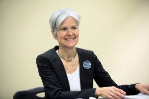 Photo of Jill Stein sitting behind a table in front of a beige wall, smiling.