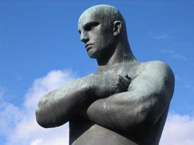 Photo of metal statue against the sky, arms crossed, torso and bald head visible.