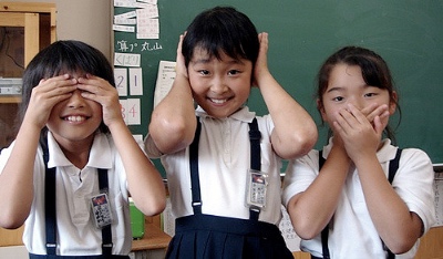 Photo of three elementary-aged schoolchildren in black and white uniforms in front of a chalkboard, covering their eyes, ears, and mouth respectively.