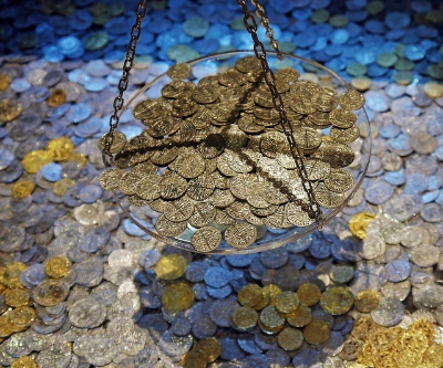 Photo of one pan of a balancing scale covered with coins against a backdrop of more coins.