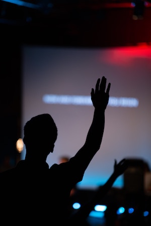 Photo: Silhouette of short-haired person with hand raised against a blurred presentation screen.