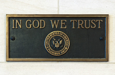 Photo of brass plaque against a white stone wall. Plaque contains seal of the United States and the words "In God We Trust".