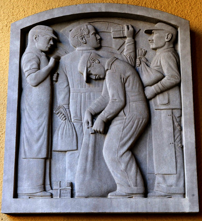 Photo of a bas relief in limestone depicting four postal workers in a frame. Three workers wear caps and aprons, holding bags. The fourth, behind them, wears a suit and holds up a package tied with string.