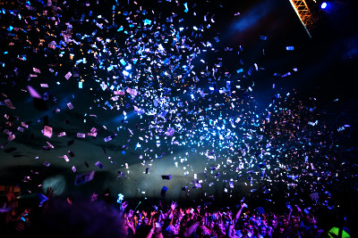 Photo of confetti being shot over an outdoor concert audience at night, catching the stage lights.