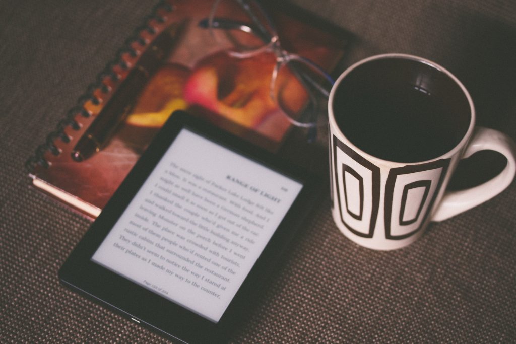 An e-reader with a cup of coffee, a notebook, a pen, and a pair of reading glasses.