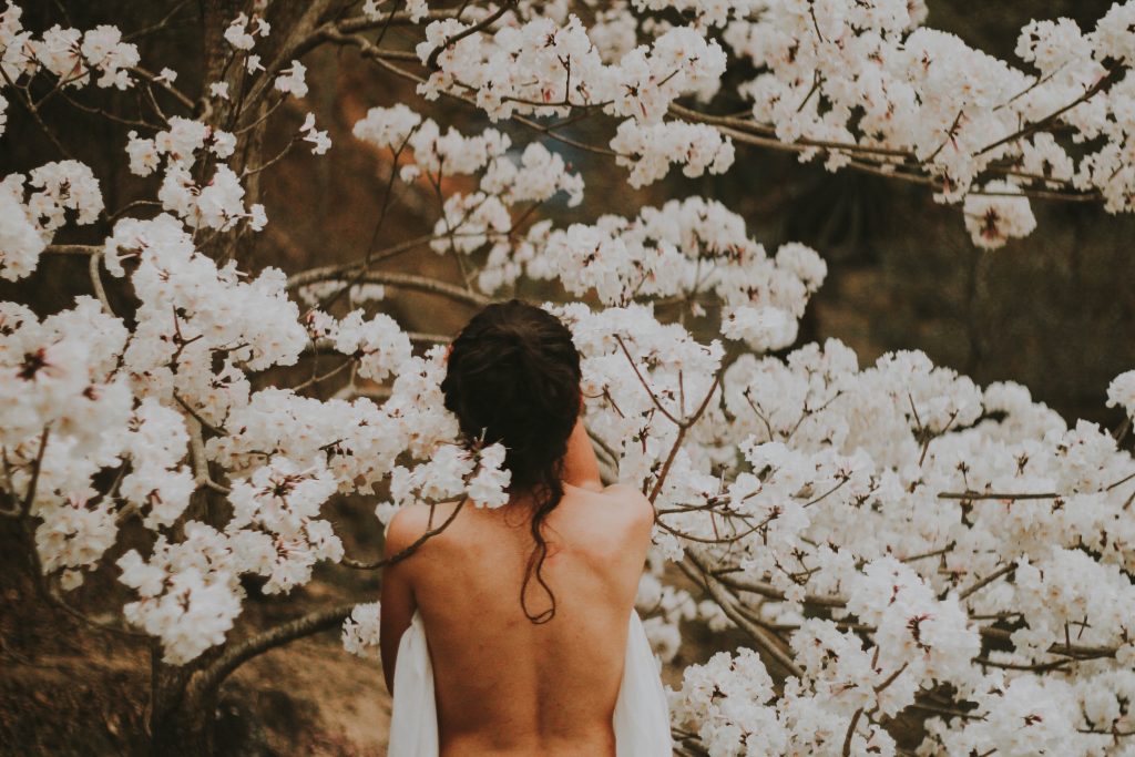 Viewed from the back and from the waist up, a nude person stands among a tree full of white flowers.