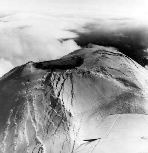 Image shows the summit of Mount St. Helens. There is a wide crater in the top, with vertical cracks radiating down through the snowcap.