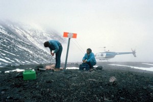 Image shows two geologists working on rocky ground. There is an orange and white reflector on a post between them. A helicopter is in the mist in the background. A portion of the mountain's flank is visible in the background, but most of the view is fogged in.