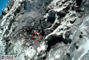 Image shows the inside of Mount St. Helens's crater. A red circle outlines David Johnston, who is a tiny figure climbing among the enormous dark gray rock walls.
