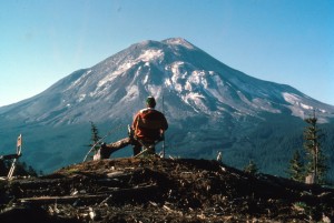 Image shows Mount St. Helens, with a man sitting in a chair on a clearcut ridge watching it.