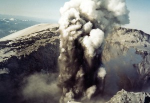Image is an aerial view of the crater, looking down into it, as a column of ash and steam rises.