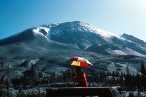 Image shows Mount St. Helens with a very good view of the Bulge, which is enormous and has changed the whole profile of the mountain. There is a geologist under a red, yellow, white, and blue striped umbrella in the foreground, keeping an eye on the volcano.