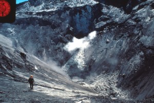 Image is looking down into Mount St. Helens's crater. There is steam rising from the crater floor. David Johnston is walking down the steep slope towards the bottom.