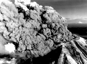 Image shows the summit of Mount St. Helens. It has been planed off, with a gap to the left, and a huge column of ash is pouring from it towards the left.