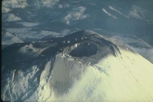 Image shows the summit of Mount St. Helens from the air. There is a large crater in the foreground, and a smaller one just behind it, a bit further down on the summit. The whole top is stained with dark gray ash.