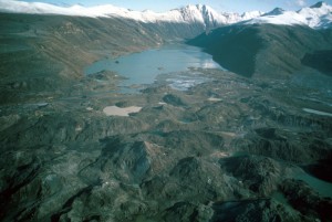 Image looks over the debris avalanche hummocks across the newborn Coldwater Lake. A snowcapped mountain range rises in the background.