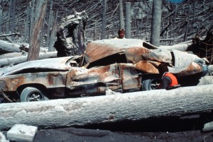 Image shows a two-door car completely destroyed by the eruption. All of the paint has been stripped off. The roof has been caved in, the windows busted out, and the body heavily dented. It's surrounded by standing trees stirpped of their limbs, and downed trees. There are men in orange vests inspecting the car.