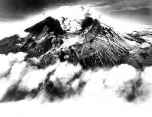 Black and white image is looking into the huge new crater of Mount St. Helens. There is an eruption plume growing from its center. The valley between the viewer and the volcano is filled with clouds of ash.