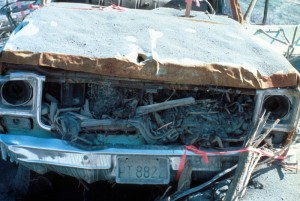 Image shows the front of a pickup truck. The grill is a mass of rubble. The hood is severely dented and covered in ash.