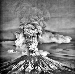 Image shows Mount St. Helens in full eruption on May 18th, 1980. A tall eruption column is barreling into the sky. To the north (center background of the photo), a long, curving, ground-hugging pale cloud runs into the distance.