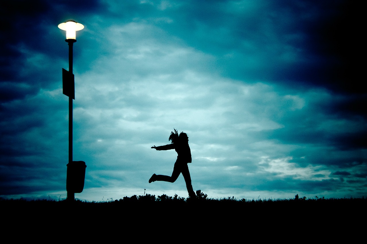 Image shows a cloudy, dark blue night sky, a lampost, and dark grassy ground. The silhouette of a running girl can be seen in the center.