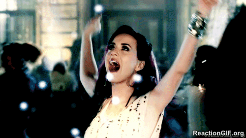 Gif shows Katy Perry dancing in a circle waving her arms while fireworks go off in the background.
